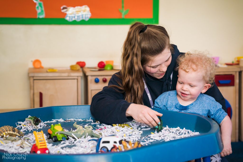 Nursery worker with child on her lap playing in a sand pit with paper and toys.